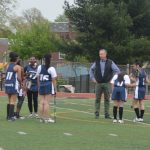 Mr. Galbraith standing with some of the 2016-2017 team, with his hands on his hips. There are five girls pictured and they are standing on the field.